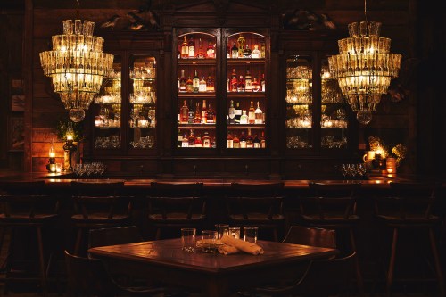 A cozy bar with a wooden counter, illuminated shelves of liquor, and elegant chandeliers creating a warm ambiance.
