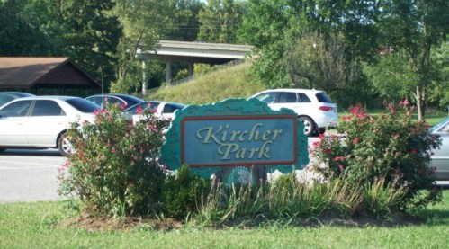 Sign for Kircher Park surrounded by flowers and parked cars, with a highway visible in the background.