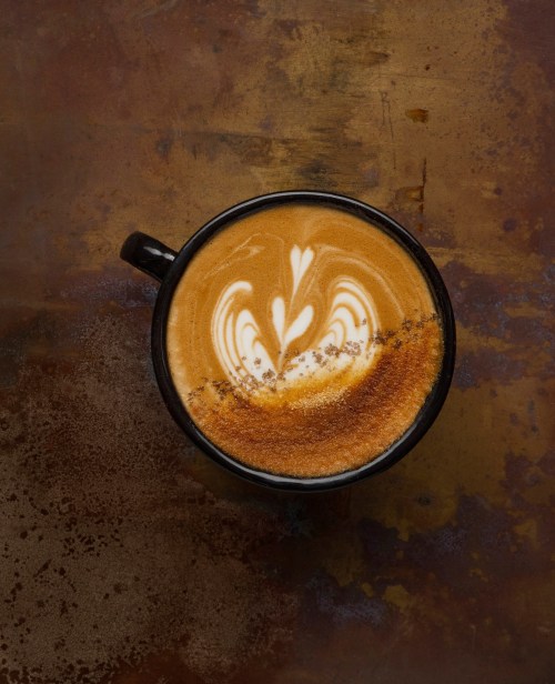 A black coffee cup with latte art on a textured, rust-colored surface.
