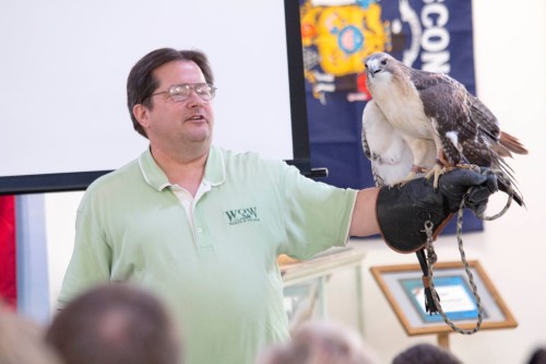 A man in a green shirt holds a hawk on his gloved hand, speaking to an audience in a classroom setting.