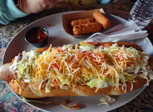 A large hot dog topped with lettuce, tomatoes, and cheese, served with mozzarella sticks and a side of salsa.