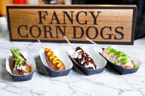 Four gourmet corn dogs with various toppings, displayed in paper trays, in front of a sign that reads "FANCY CORN DOGS."