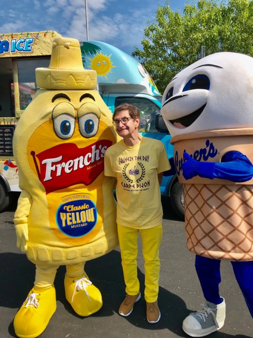 A person in yellow stands between a mustard mascot and an ice cream cone mascot at a food event.