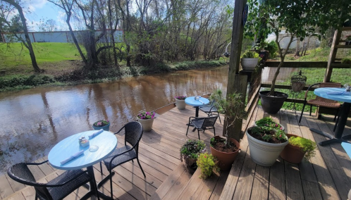 A cozy outdoor dining area by a calm river, featuring tables, chairs, and potted plants under a sunny sky.