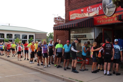 A long line of cyclists in colorful jerseys waiting outside a soda fountain shop on a sunny day.