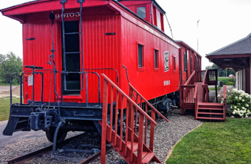 A red caboose on tracks, with wooden steps leading up to it, surrounded by grass and a building in the background.