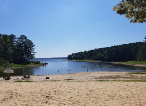 A sandy beach by a calm lake, surrounded by trees, with people enjoying the water and colorful umbrellas in the foreground.