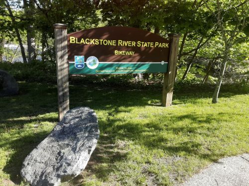 Sign for Blackstone River State Park Bikeway, surrounded by greenery and a large rock.