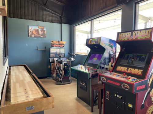A game room featuring arcade machines, including Mortal Kombat, and a shuffleboard table by large windows.