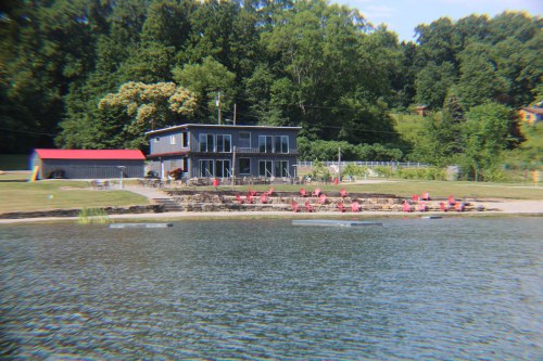 A lakeside view of a modern house with a red roof, surrounded by green trees and chairs on the shore.