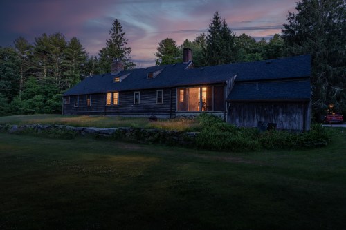 A rustic wooden house at dusk, with warm lights glowing from the windows, surrounded by trees and a grassy lawn.