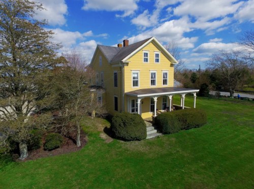A yellow two-story house with a porch, surrounded by green grass and trees under a blue sky with fluffy clouds.