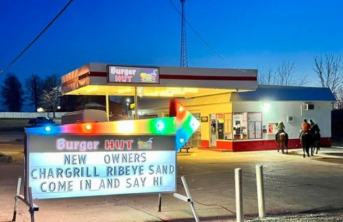 A brightly lit Burger Hut with a sign announcing new owners and a special ribeye sandwich, set against a twilight sky.