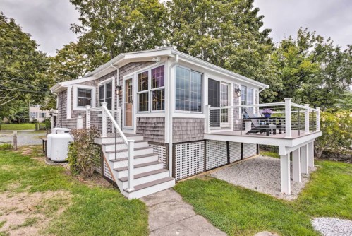 A charming cottage with a deck, surrounded by greenery and a gravel path, under a cloudy sky.