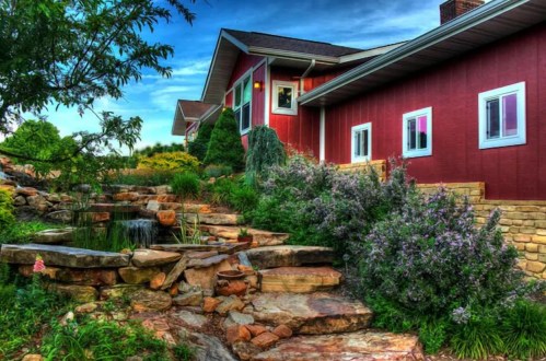A red house with colorful windows surrounded by lush greenery and a stone pathway leading to a small pond.