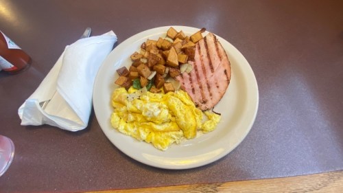 A plate of scrambled eggs, grilled ham, and seasoned potato cubes on a brown table.