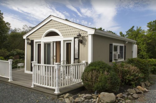 A small, modern house with a porch, surrounded by greenery and rocks, under a partly cloudy sky.