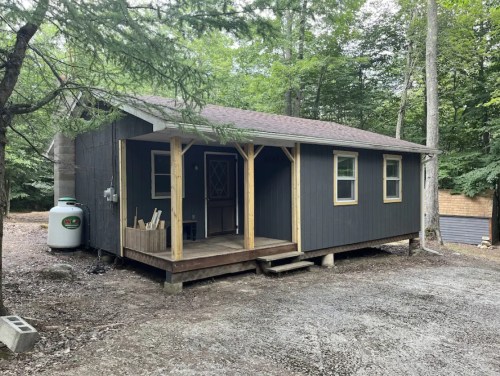 A small, dark-colored cabin with a porch, surrounded by trees and gravel, in a wooded area.