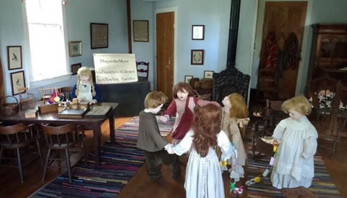 A vintage classroom scene with dolls dressed in period clothing, interacting around a wooden table and colorful rug.