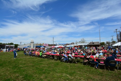 A large crowd gathers at an outdoor event with tables set up under a blue sky and scattered clouds.