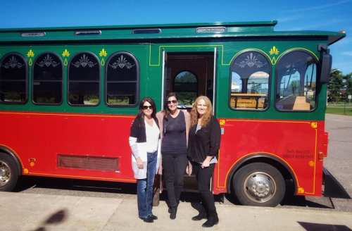 Three women stand in front of a colorful trolley bus, smiling on a sunny day.