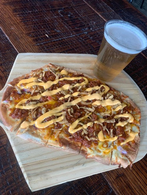 A slice of pizza topped with sauce, cheese, and drizzled with yellow sauce, alongside a glass of beer on a wooden board.