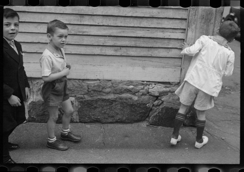 Three young boys are playing outside near a wooden wall, with one boy looking back and two others engaged in different activities.