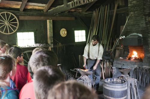 A blacksmith demonstrates his craft in a historic workshop, with an audience watching and tools in the background.