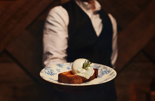 A waiter presents a plate with a slice of cake topped with a scoop of ice cream and a sprig of rosemary.