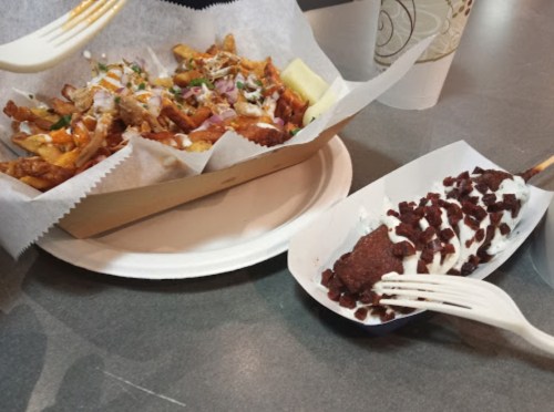 A plate of loaded fries topped with cheese, bacon, and green onions, alongside a chocolate-covered dessert on a stick.
