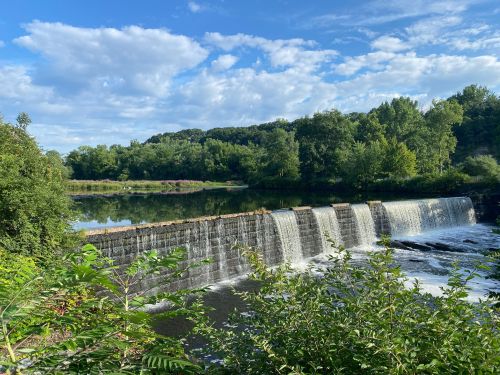 A serene waterfall cascades over a stone dam, surrounded by lush greenery and a clear blue sky.