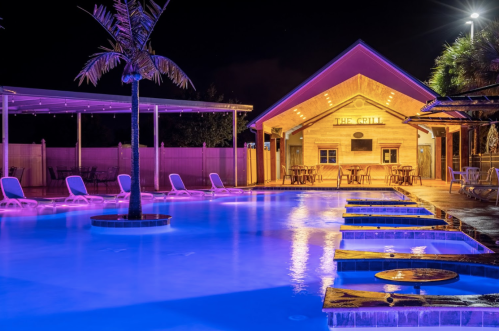 A beautifully lit pool area at night, featuring lounge chairs, palm trees, and a wooden grill house in the background.