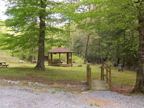 A serene outdoor area with a gazebo, picnic tables, and a wooden bridge surrounded by trees.