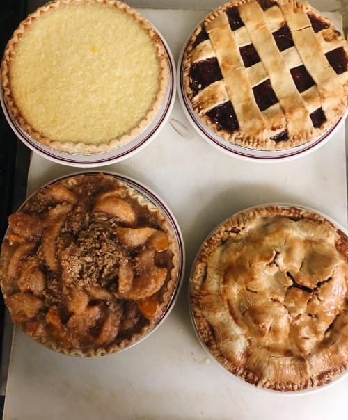 Four freshly baked pies on a countertop: lemon, cherry lattice, apple crumble, and a golden brown pie.