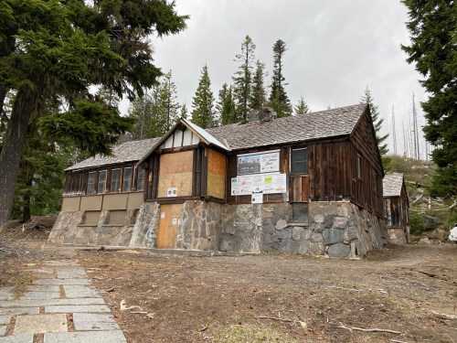 A rustic wooden building with a stone foundation, surrounded by trees and a gravel path, under a cloudy sky.