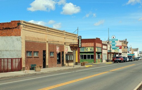A small town street featuring vintage storefronts, a café, and parked cars under a blue sky with scattered clouds.