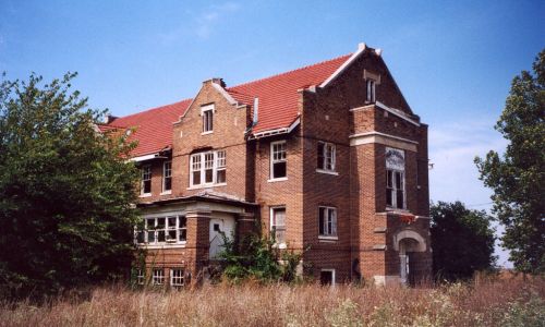 Abandoned brick building with a red roof, surrounded by overgrown grass and trees under a clear blue sky.