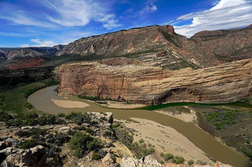 A scenic view of a winding river surrounded by rugged cliffs and green vegetation under a blue sky with clouds.
