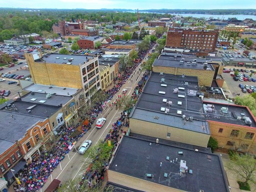 Aerial view of a bustling street parade with crowds, colorful umbrellas, and buildings lining the road.