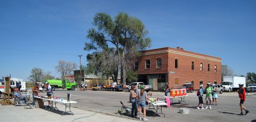 A small town street scene with people at tables, a brick building, and vehicles under a clear blue sky.