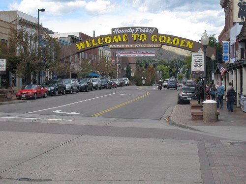 A street view of Golden, Colorado, featuring a welcoming arch and parked cars along a tree-lined road.