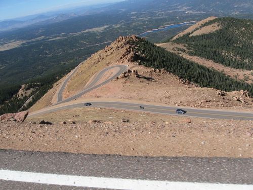 Winding road curves around a mountain peak, with a scenic view of valleys and lakes in the background.