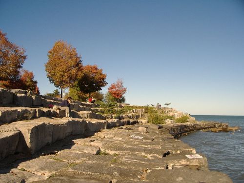 A rocky shoreline with autumn trees and people enjoying the view by the water under a clear blue sky.
