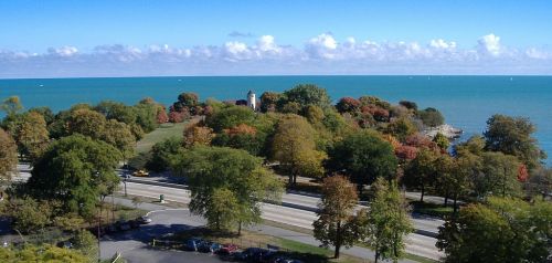 A scenic view of a park with colorful trees by a lake under a clear blue sky and fluffy clouds.