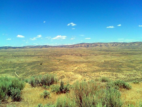 A vast, arid landscape with rolling hills, sparse vegetation, and a clear blue sky dotted with clouds.