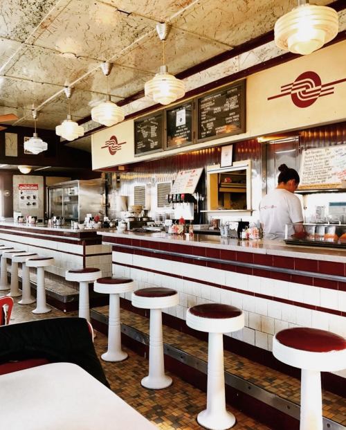 A retro diner interior with white and red stools, a counter, and a staff member preparing food.