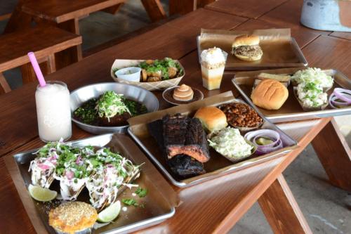 A table filled with various dishes, including tacos, burgers, sides, and drinks, all served on metal trays.
