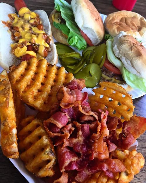 A plate of waffle fries topped with bacon, alongside burgers, hot dogs with cheese, and pickles.
