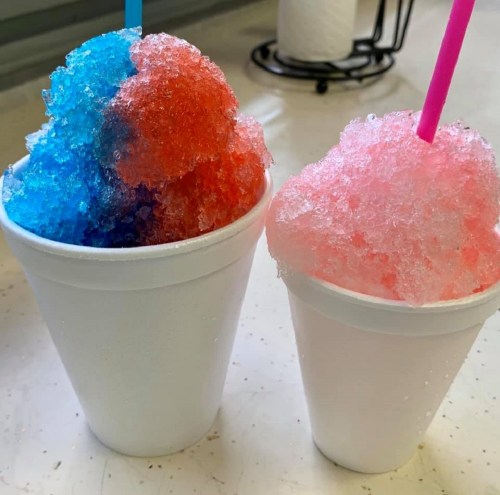 Two cups of colorful shaved ice, one blue and red, the other pink, each with a straw, on a table.