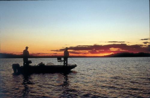 Two fishermen silhouette on a boat at sunset, with vibrant colors reflecting on the calm water.